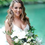 A smiling woman in a white wedding dress holds a bouquet of white flowers, standing in front of a green backdrop.